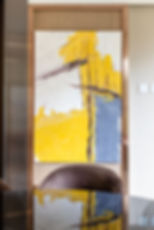 Abstract painting with bold yellow, white, and gray brushstrokes. Framed and displayed in a modern room with a brown chair below. MAD Studio