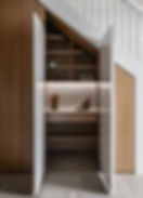 Open wooden cabinet under stairs with shelves, kitchenware, and books. Warm lighting creates a cozy and organized atmosphere. MAD studio