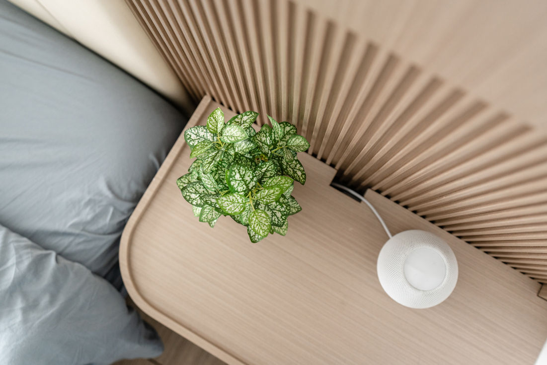 Modern minimalist bedroom interior with a wooden nightstand featuring a vibrant green plant and a sleek smart speaker. The neutral tones and clean lines exemplify contemporary interior design trends, perfect for creating a serene and stylish living space. 