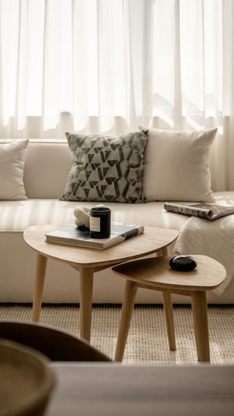 A cozy living room featuring a neutral-toned sofa adorned with patterned and plain cushions. In front of the sofa, there are two nested wooden coffee tables. The larger table holds a book and a dark candle, while the smaller table has a decorative stone. A soft beige throw rests on the sofa, complementing the warm, inviting ambiance created by the sheer curtains filtering natural light.