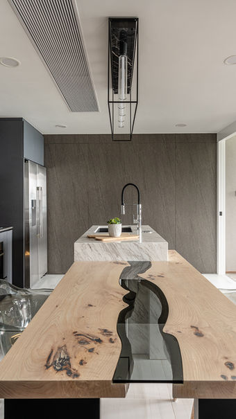 Modern kitchen and dining area with sleek, gray cabinetry and a marble backsplash. A unique wooden dining table with a glass river design serves as a centerpiece, paired with transparent chairs. A large stainless steel refrigerator and a stylish island with a black faucet complete the contemporary look. The space is open and bright, featuring a sliding glass door leading to a cozy living area with a leather sofa, Residential interior design