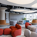 School lounge area with unique, rounded seating in shades of gray and red. The space features minimalist design elements, including a large circular light fixture and neutral geometric carpeting. Stylish chairs are arranged around pillar accents with warm-toned cushions, complementing the contemporary aesthetic. Large windows allow natural light, highlighting the clean lines and open layout.