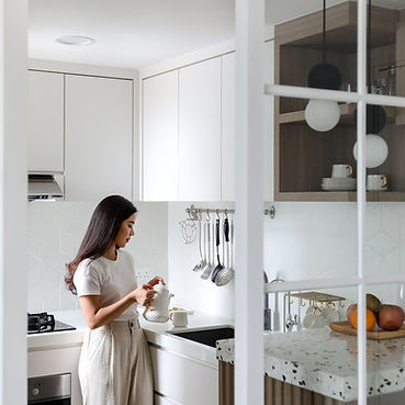 A woman in a white shirt and beige pants is standing in a modern kitchen, pouring liquid from a kettle into a cup. The kitchen features white cabinets, a white counter, and a tile backsplash. Various utensils hang on the wall, and a bowl of fruit sits on a nearby counter. The scene is viewed through a glass-framed partition.